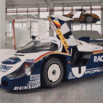 white and blue Porsche race car with doors open upwards inside of a warehouse with lots of windows in background