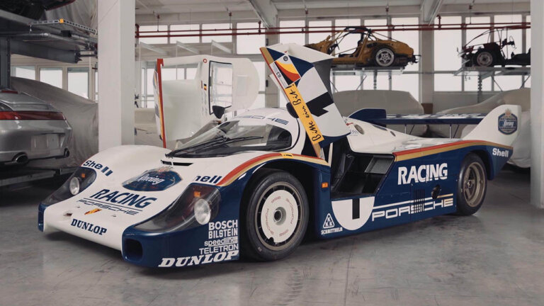 white and blue Porsche race car with doors open upwards inside of a warehouse with lots of windows in background