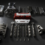 Engine parts layed out on a dark concrete floor. Metal cam shafts, manifolds, pistons, next to red valve cover