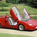 red Lamborghini Countach with scissor doors open and wood fence running behind it. Large grassy field behind both of those objects
