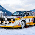 White Audi Sport Quattro S1 E2 with yellow stripes parked with lights on in the snow with mountains behind it