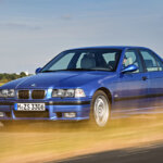 Blue m3 e36 on road near a grass field. Grass infront of the car is passing it really fast