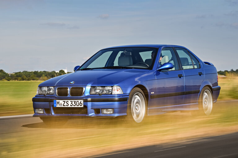 Blue m3 e36 on road near a grass field. Grass infront of the car is passing it really fast