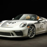 Silver Porsche 911 with the number 49 in a white circle. Car in a dark room with black background