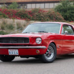 Red 1964 Ford Mustang parked infront of a curb and a building is in the background