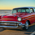 Red Chevy Nomad station wagon pictured on the side of a freeway with the ocean and sunset behind it