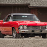Red and black Chevy El Camino parked on gravel and in front of a red building
