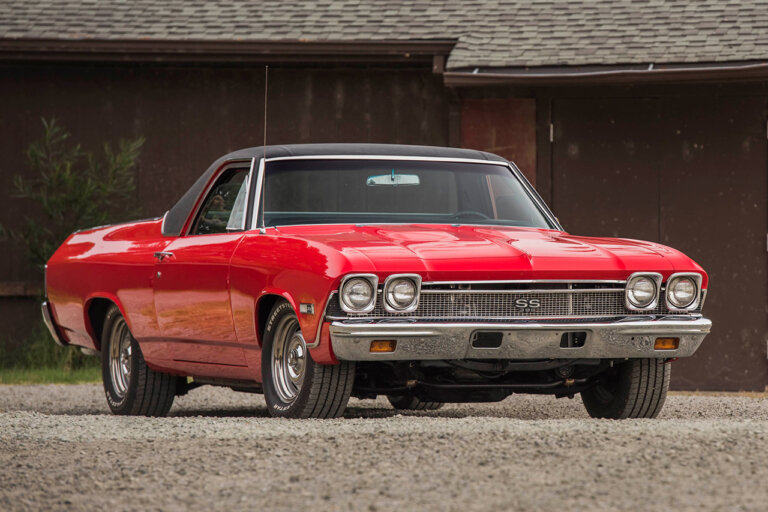 Red and black Chevy El Camino parked on gravel and in front of a red building