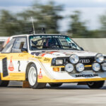 Yellow and white Audi Quattro with a man in a race suit driving past trees