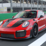 Red and black Porsche 911 GT2RS with a man driving on a race track with seats in background