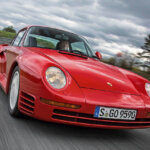 Red Porsche 959 driving on an asphalt road with grey clouds in background