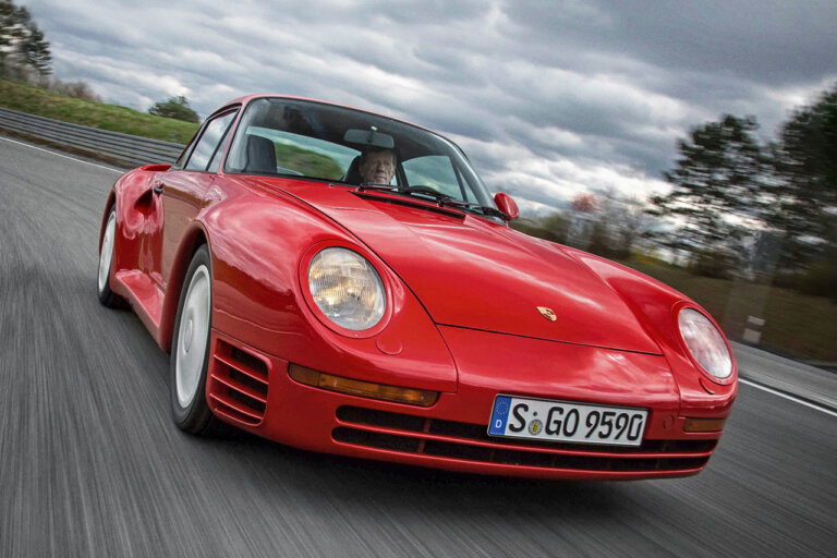 Red Porsche 959 driving on an asphalt road with grey clouds in background