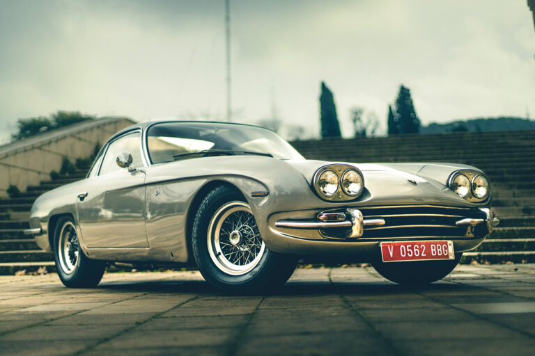 Silver Lamborghini 350 GT car parked on stone ground with trees and a building behind it