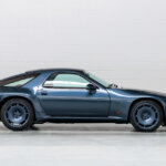 Blue Porsche 928 parked sideways against a white brick wall