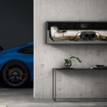 Photo of a blue Porsche 911 behind a cement wall with an exhaust hanging above a glass table