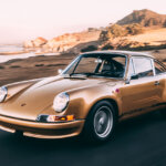 Gold Porsche 911 driving along the coastline with hills in background