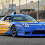 Blue and yellow Honda NSX parked on racetrack with fence in the background