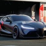 Dark grey Toyota Supra with a large red and white decal towards the rear of the car as the car is staged outside of a garage