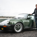 Man in jacket and jeans standing next to a green Porsche 930 with cloudy skies in background