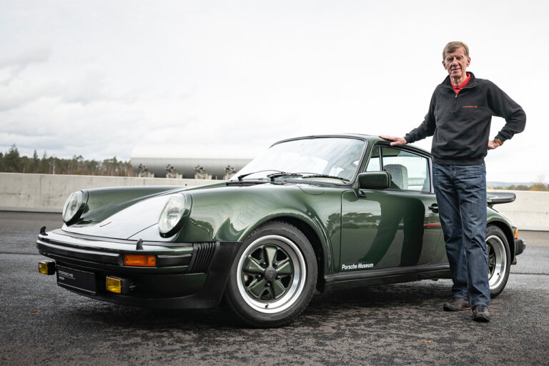 Man in jacket and jeans standing next to a green Porsche 930 with cloudy skies in background