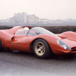 Red Ferrari 330 P4 racecar with a man wearing a white helmet in the driver seat with buildings in the background