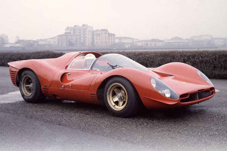 Red Ferrari 330 P4 racecar with a man wearing a white helmet in the driver seat with buildings in the background