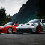Dark photo of two Porsches parked next to one another, one silver and the other red with trees and overcast sky in background