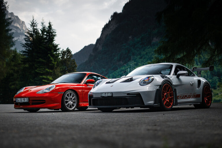 Dark photo of two Porsches parked next to one another, one silver and the other red with trees and overcast sky in background