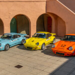 Blue, green, orange and yellow Porsche 911's parked next to one another in a courtyard surrounded by an orange building