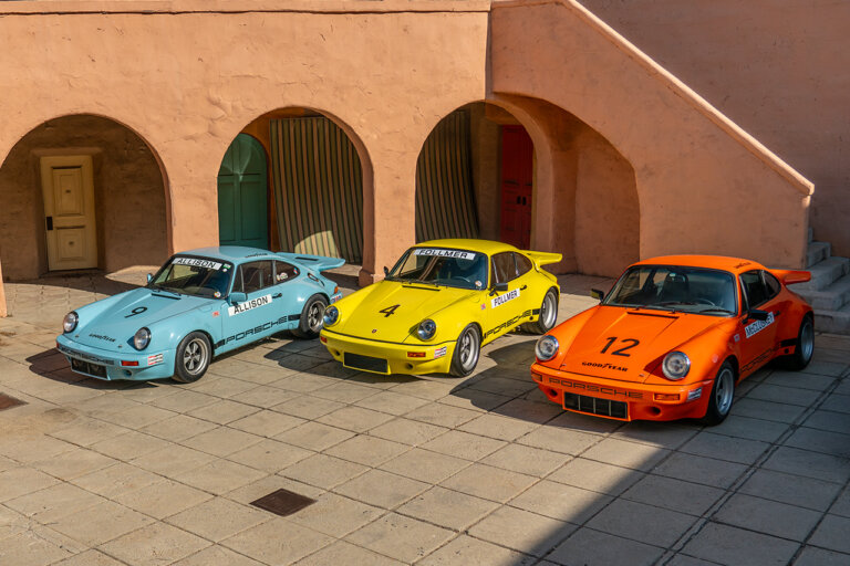 Blue, green, orange and yellow Porsche 911's parked next to one another in a courtyard surrounded by an orange building
