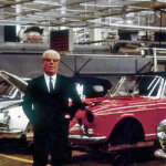 Man in a suit with white hair and glasses standing next to several cars ready to be worked on in a warehouse