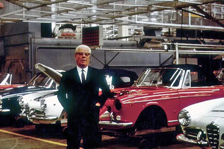 Man in a suit with white hair and glasses standing next to several cars ready to be worked on in a warehouse