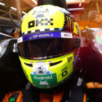 Race car driver with a green helmet covered in stickers strapped down with a Sparco racing harness with people on either side of the driver cockpit