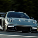 Silver RML Group P39 race car driving on a race track with a man wearing a white helmet with trees in background