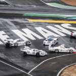 White text saying Le Mans in all caps behind Porsche race cars staged on a bank of a wet race track.