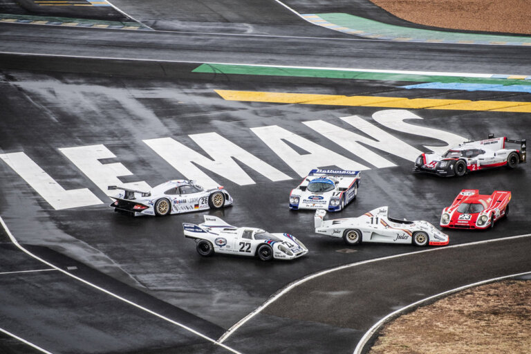 White text saying Le Mans in all caps behind Porsche race cars staged on a bank of a wet race track.
