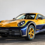 Yellow and blue Porsche 911 Dakar with yellow rims parked in a room with square concrete floor and a white wall.