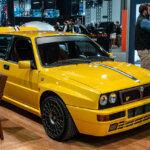 Yellow Lancia Delta Evo II parked on wood floor next to leather chairs inside of the SEMA 2024 exhibit