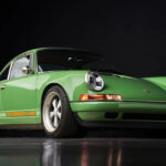 Green with orange accents of a Singer Porsche 911 pictured in a dark room with reflective floor and brick wall in background.