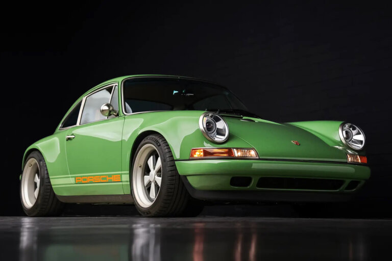 Green with orange accents of a Singer Porsche 911 pictured in a dark room with reflective floor and brick wall in background.