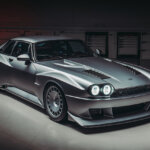 Silver car with 2 lights in both headlights parked in a dark room with reflective floor and red taillight projecting red on the background.