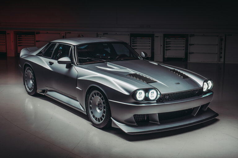 Silver car with 2 lights in both headlights parked in a dark room with reflective floor and red taillight projecting red on the background.
