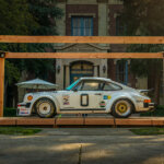 White Porsche 911 930 with stickers and racing decals and the number 0 in black on the driver door. The car is on a wooden platform resembling a box that sits in a grassy courtyard with a tall building in the background