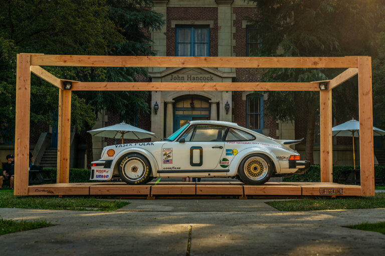 White Porsche 911 930 with stickers and racing decals and the number 0 in black on the driver door. The car is on a wooden platform resembling a box that sits in a grassy courtyard with a tall building in the background
