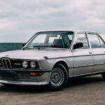 Silver BMW E28 M535i sedan parked at an angle with sun setting behind clouds in the sky in the background.
