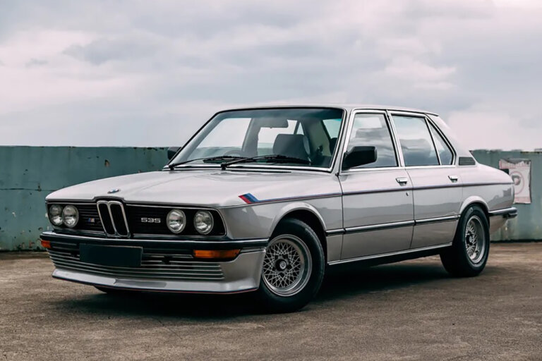 Silver BMW E28 M535i sedan parked at an angle with sun setting behind clouds in the sky in the background.