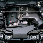 BMW S54 engine with carbon fiber valve covers and intake manifold with a strut bar going across the top attached to each tower inside of a silver car.