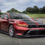 Red Ford Mustang GTD race car parked at an angle on a racetrack with trees, grass and cloudy sky in the background.
