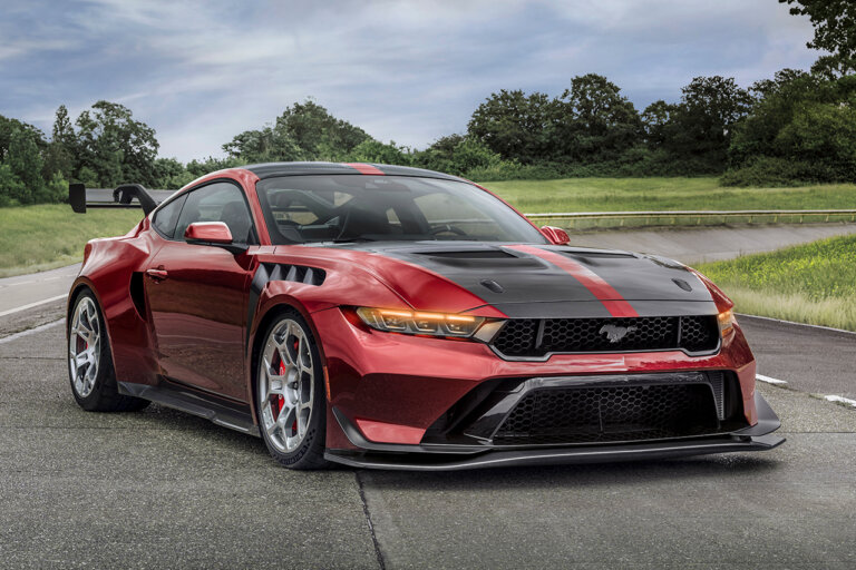 Red Ford Mustang GTD race car parked at an angle on a racetrack with trees, grass and cloudy sky in the background.