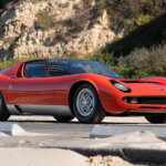 Red Lamborghini Miura P400 parked on a road next to sand in the foreground and a cliffside in the background.